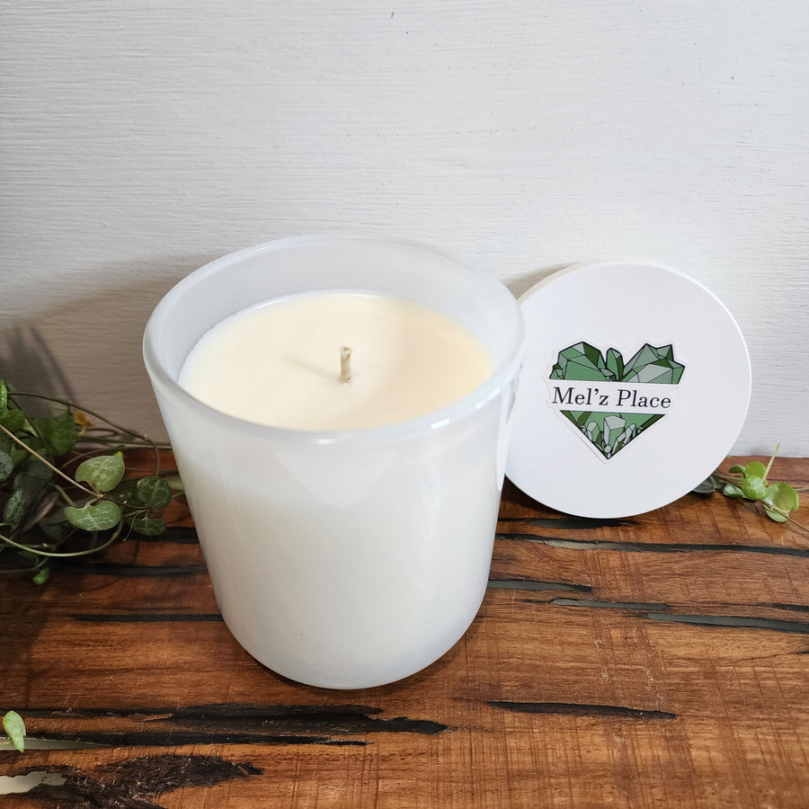 A white soy candle with a clear container and a branded label that reads 'mel's place' with a green heart and leaf design. The candle is placed on a wooden surface with a white background.