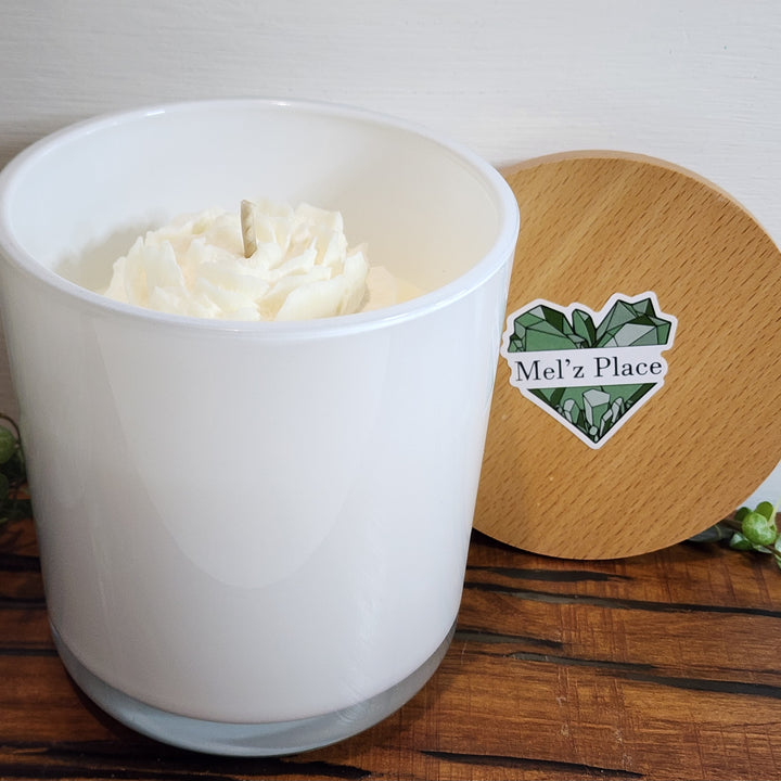 A white soy candle is shown in an opaque container with a wooden lid next to it. The candle wax is white with a flower detail in the wax.