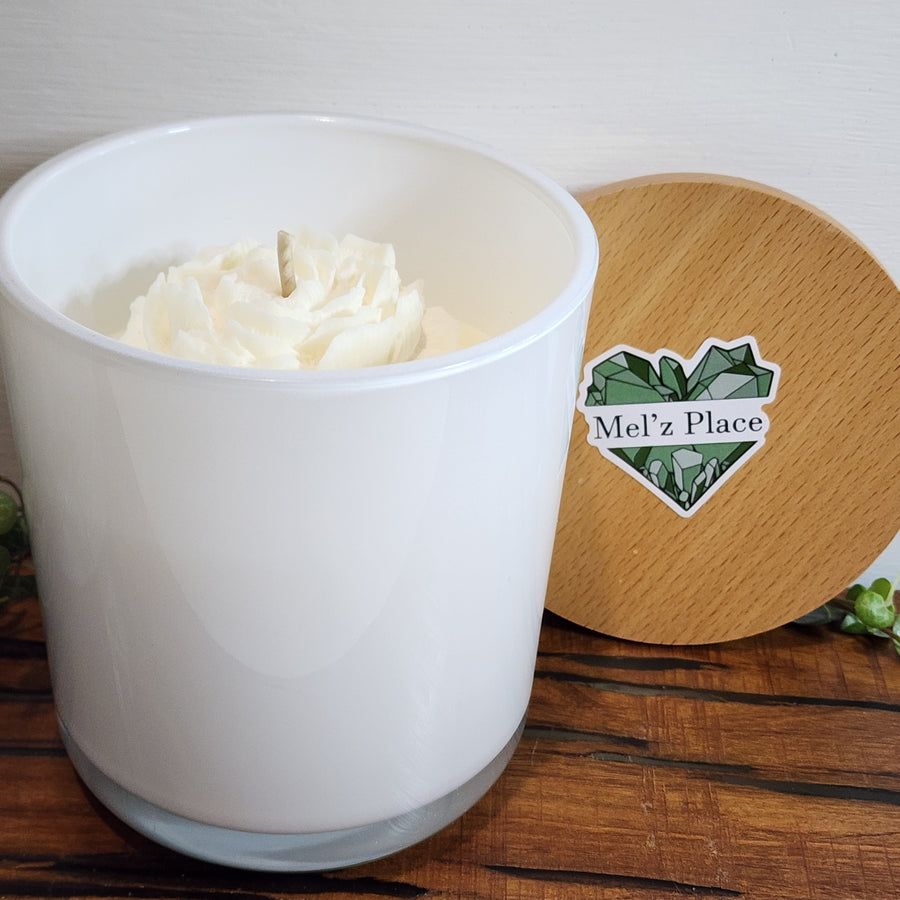 A white soy candle is shown in an opaque container with a wooden lid next to it. The candle wax is white with a flower detail in the wax.